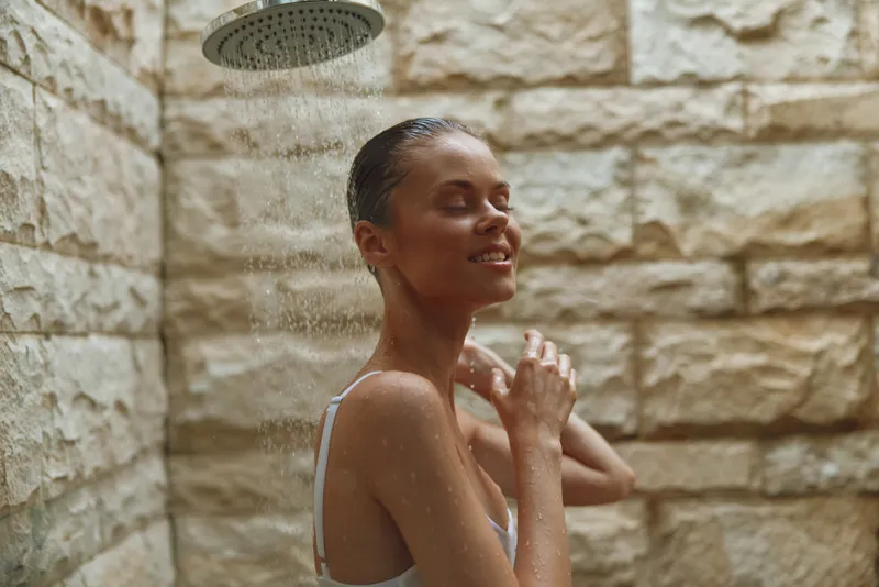 woman enjoying outdoor shower with stone wall background, wet hair, natural light, peaceful expression, summer wellness and relaxation moment