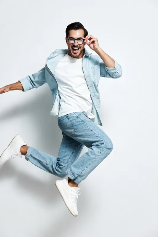 Full body joyful happy young man wearing casual blue shirt and jeans jumping playfully while smiling at the camera isolated on clean white background. Lifestyle concept