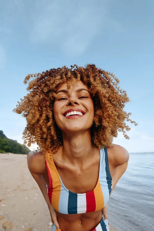 Smiling woman with curly hair wearing colorful striped swimsuit on the beach. Sunny day with clear sky and calm sea in the background. Concept of summer, happiness, and vacation.