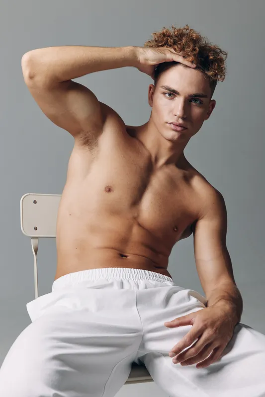 Young man with curly hair posing shirtless sitting on chair with hand on forehead, wearing white pants against gray background in studio portrait.