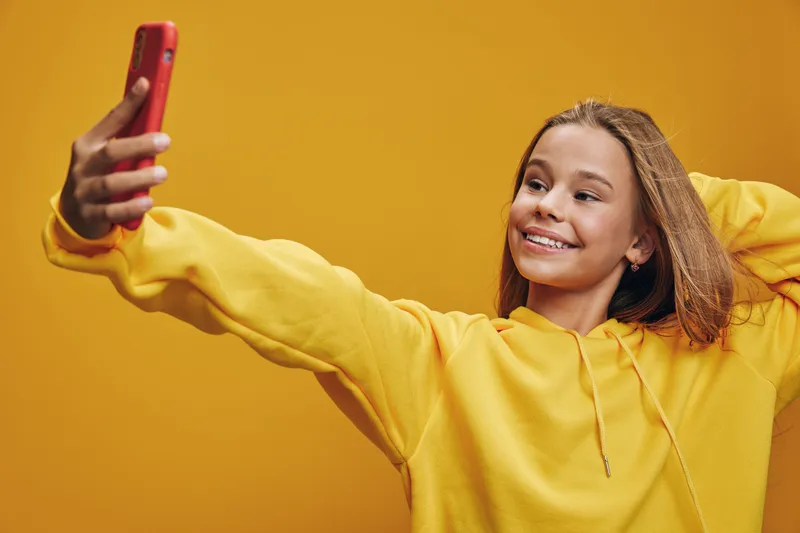 Teenage girl smiling and taking selfie photo with smartphone in yellow hoodie on bright background. Casual style and happy expression with long hair for youth technology and social media.