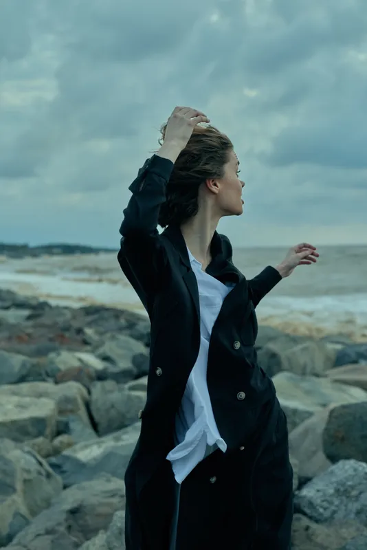 Wind swept coat and flowing white shirt by a rocky shoreline, motion in the sleeves as clouds roll over the sea, outdoor coastal scene with rugged rocks and sea spray under a moody sky