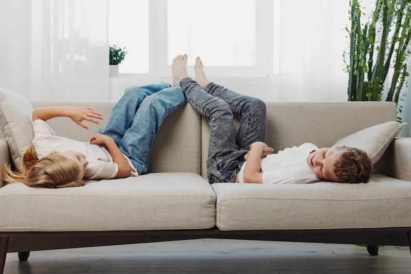 Two children relax on a beige sofa with their feet touching, in a bright living room with large windows and green plants. The scene shows comfort, playfulness, and sibling bonding.