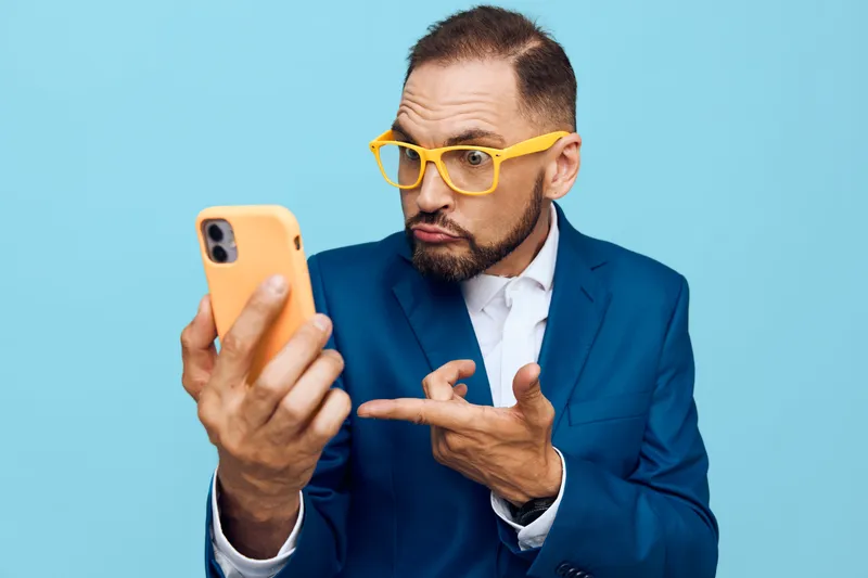 A professional business man in a blue suit on an isolated blue background, expressing curiosity and intrigue while using a smartphone.