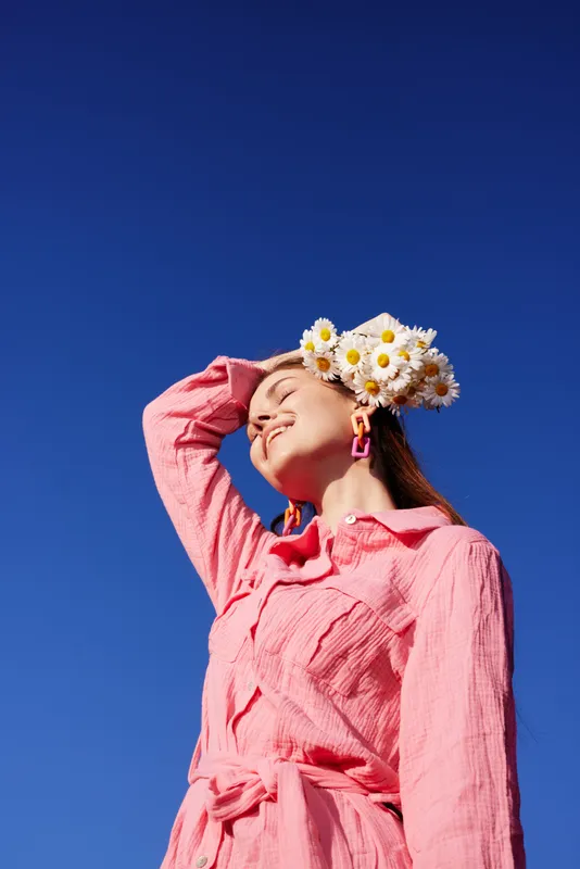 Young woman with a floral crown in a pink dress against a clear blue sky, radiating joy and confidence while embodying spring fashion and vibrant colors