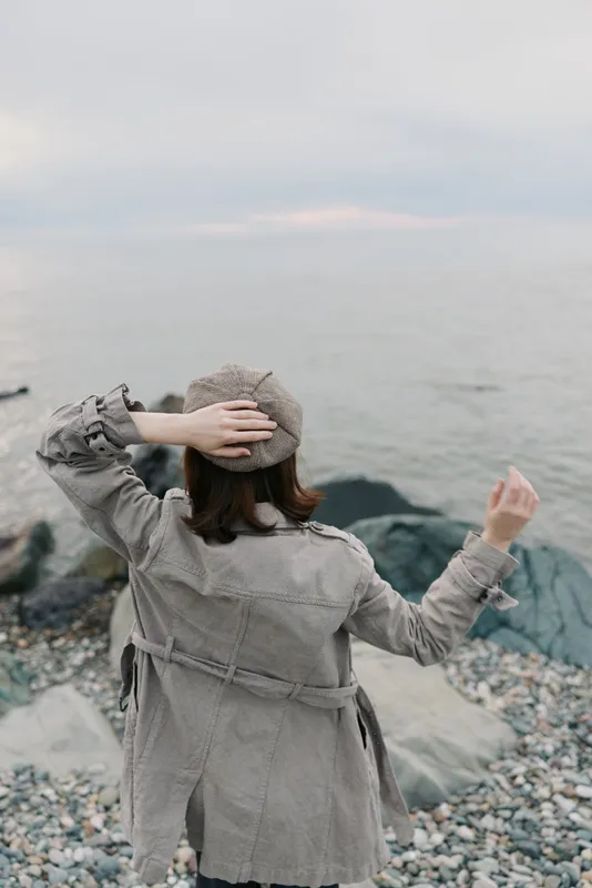 Woman in grey coat and knitted hat stands on rocky shore, looking at calm sea horizon. Concept of solitude, nature, cold weather, and peaceful coastal scenery in autumn or winter.