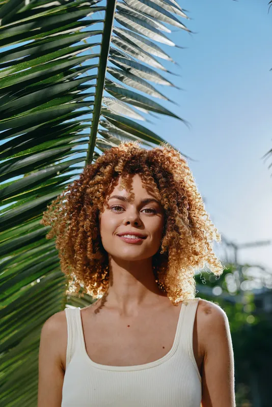 smiling woman with curly hair standing outdoors under palm leaves, wearing a white tank top with bright natural sunlight and blue sky background