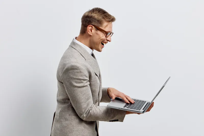 Professional man with glasses in gray suit holding a laptop and smiling, presenting a friendly, modern, corporate look. Concept: business success, technology, communication, confident professional.