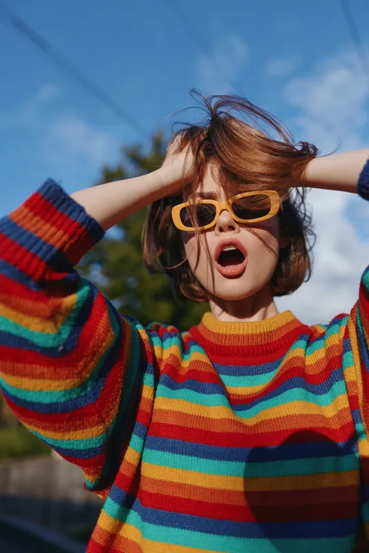 Woman portrait wearing sunglasses and colorful sweater, surprised expression outdoors; fashion short hair, rainbow stripes and sunlight on face for vibrant candid street style.