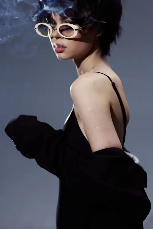 Stylish woman with short blue hair wearing sunglasses and black outfit posing with smoky background in moody light