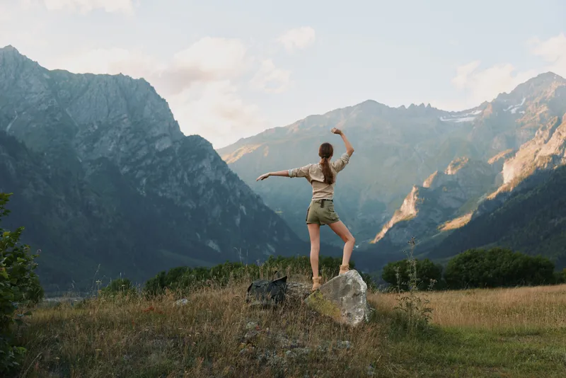Young woman stands on a rock with arms raised in a rugged valley, rocky mountains in the background and a clear sky above, conveying freedom and exhilaration.