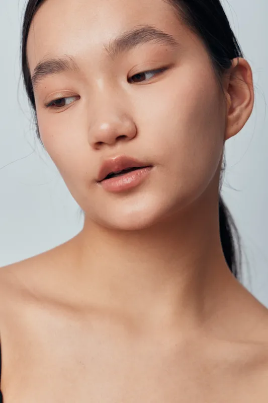Portrait of young woman with natural makeup and smooth skin looking down with closed lips and serene expression on light background. Beauty, closeup and skincare concept with bare shoulders.