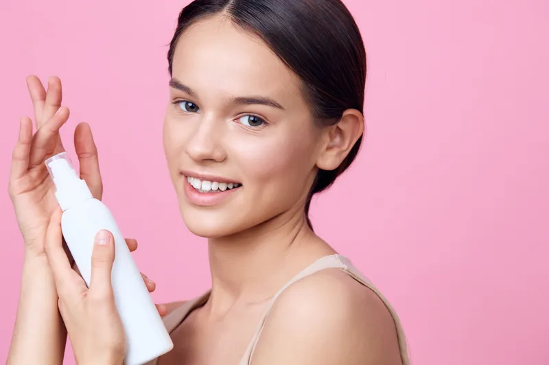 Young woman with a radiant smile holding a skincare bottle against a pink background, promoting beauty and skincare routines for healthy skin