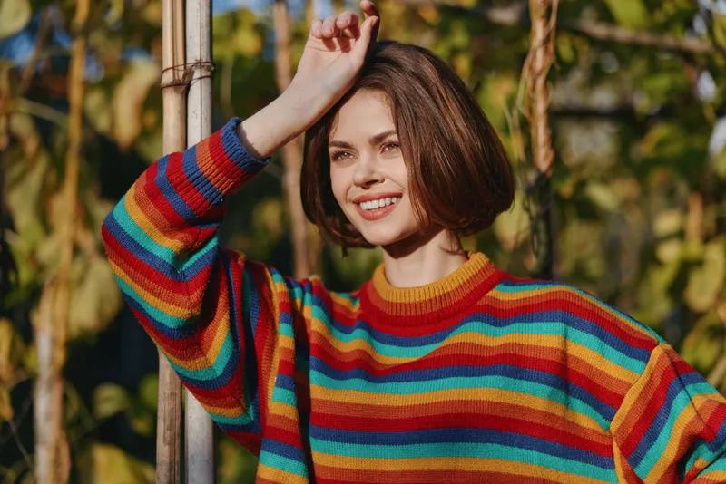 Woman smiling in rainbow sweater outdoors, portrait of young female with short hair leaning on pole, natural sunlight and autumn leaves background, casual colorful fashion, relaxed pose.