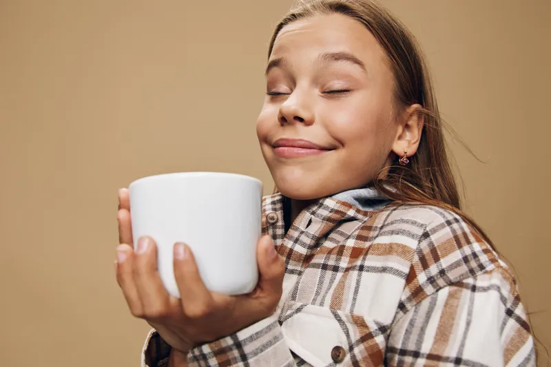 Girl with closed eyes holding white cup and smiling in cozy plaid jacket. Relaxed young female enjoying hot beverage and comfort indoors with peaceful expression on beige background.