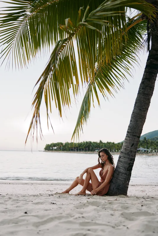 Relaxed woman sitting under palm tree on tropical beach during sunset with calm sea and serene atmosphere.