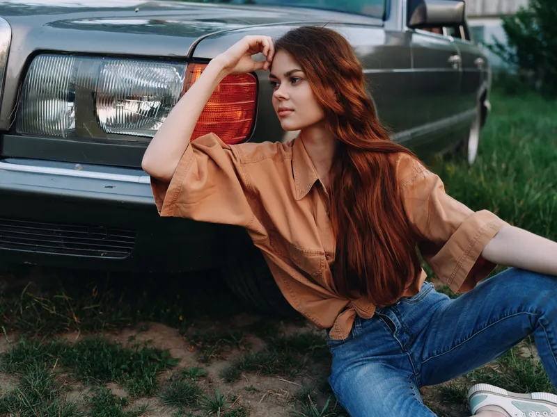 Young woman in casual outfit sitting by a vintage car, expressing thoughtful emotion, captured in warm earthy tones during golden hour.