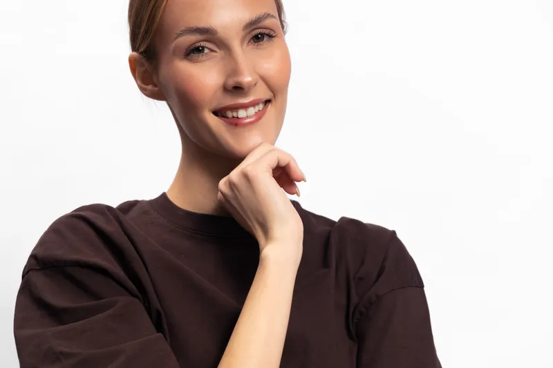Smiling young woman with clear skin and brown shirt poses with hand near chin against white background. Concept of confidence, beauty, and natural portrait with positive expression.