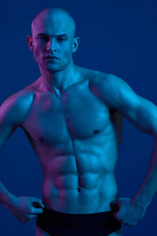 Powerful man showcasing muscular physique under vibrant blue and purple lighting, exuding confidence and strength in a captivating atmosphere.