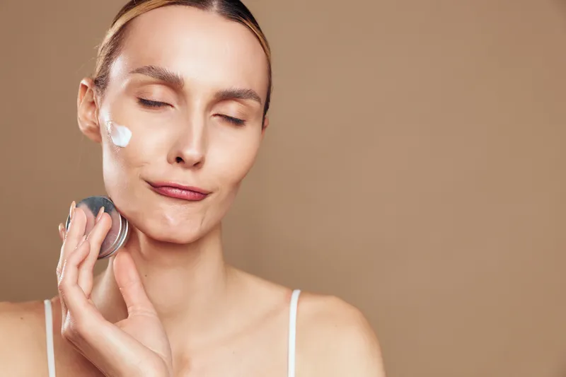 Young woman applying facial cream on cheek with cotton pad, closed eyes, neutral expression, smooth skin, beige background, skincare routine, personal care concept