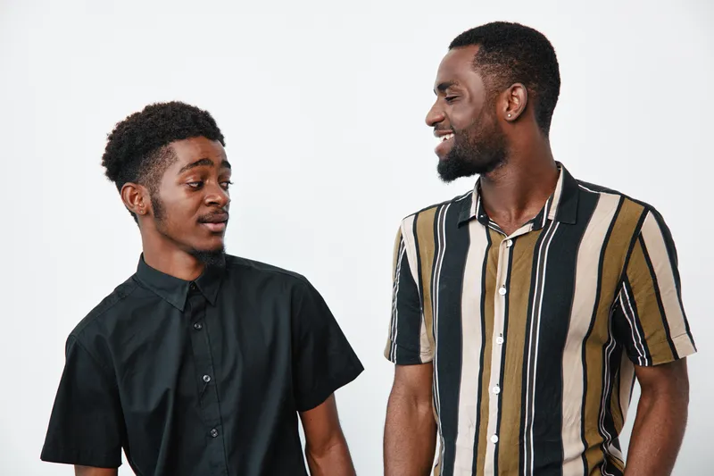 Two African American men with dark skin standing and smiling, casual clothing, one in striped shirt and the other in black shirt, friendly interaction on white background.
