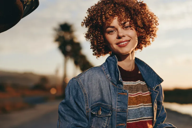 Woman lifestyle outdoors, smiling with relaxed posture, curly hair and denim jacket in golden sunset light, soft breeze feel, scenic road background, warm natural colors, evening glow sweet
