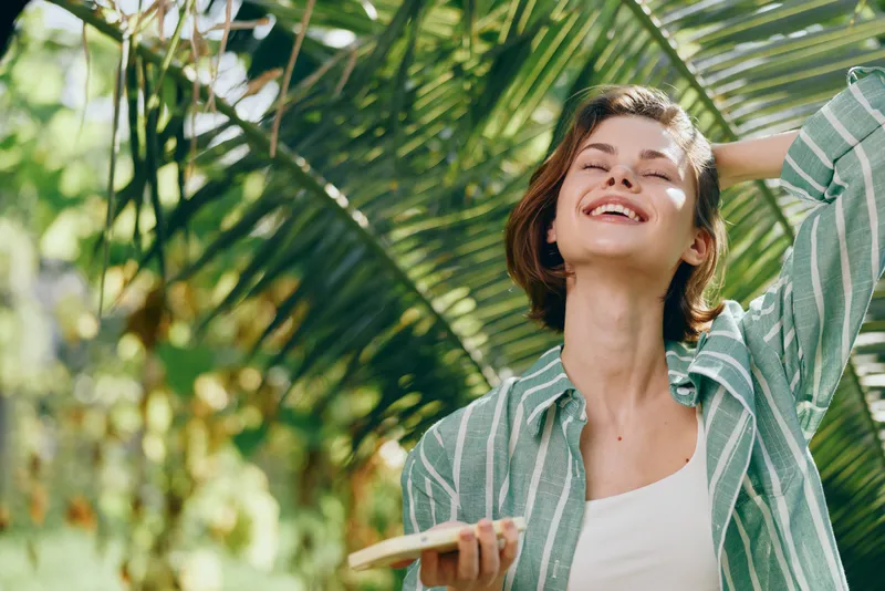 Portrait of a smiling woman holding a phone, bathed in sunshine amid lush tropical foliage.