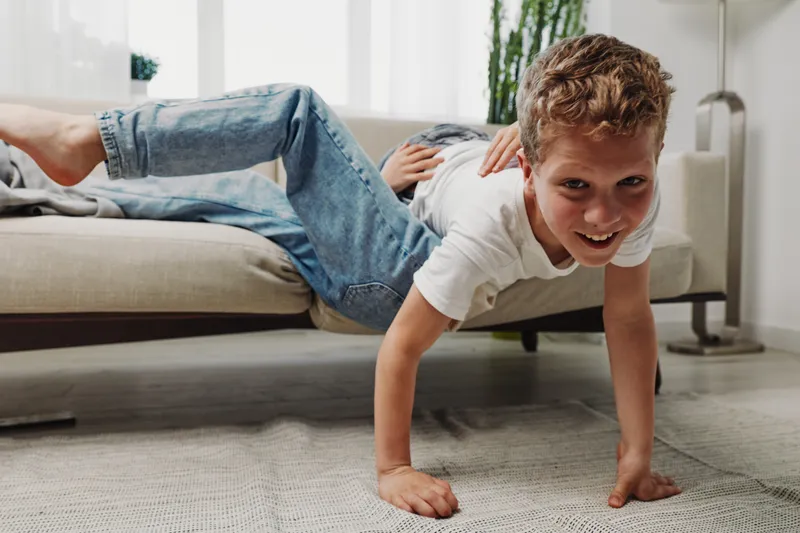 Happy boy in casual clothes balancing on hands with legs on couch in bright living room. Playful child having fun indoors with active pose and smile.
