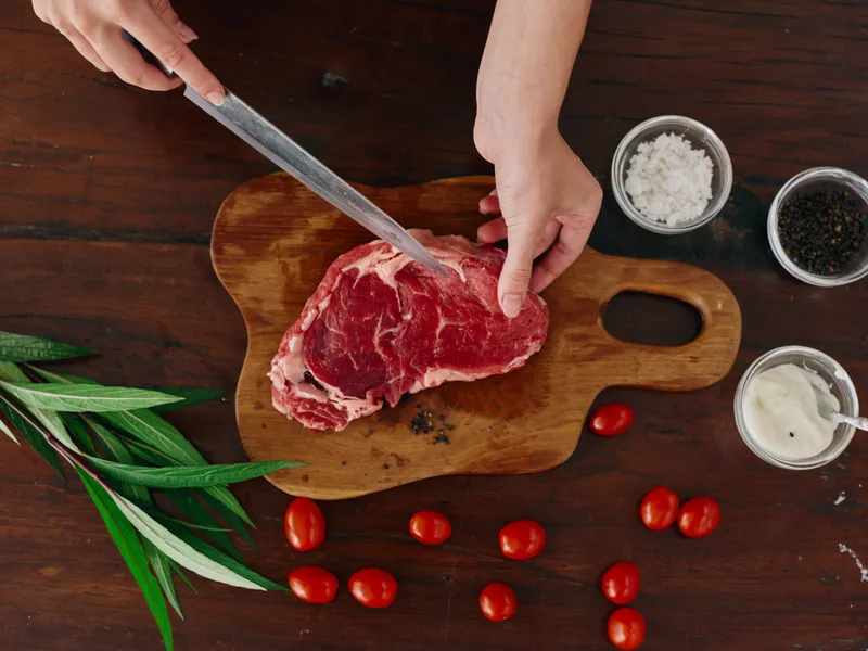 Женщина с ножом в руке нарезает свежее мясо стейка для жарки на кухне