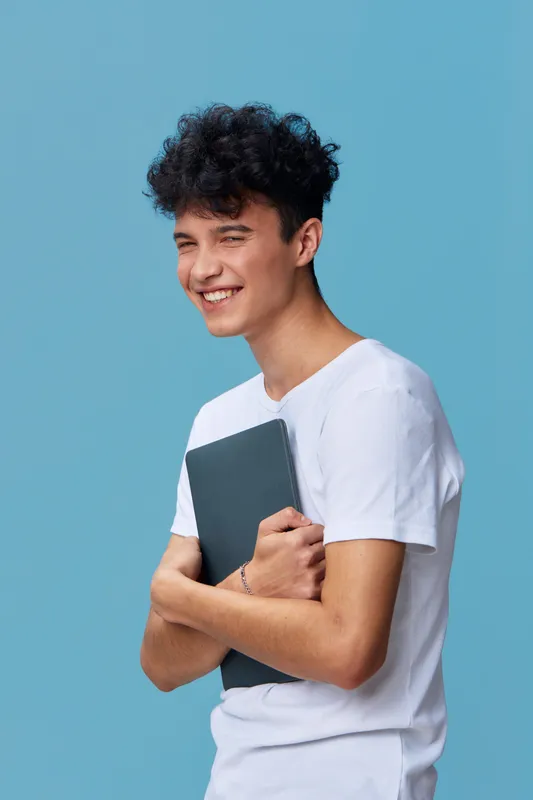Young smiling man wearing a white t-shirt holding a notebook, standing against a blue background, expressing happiness and casual style.