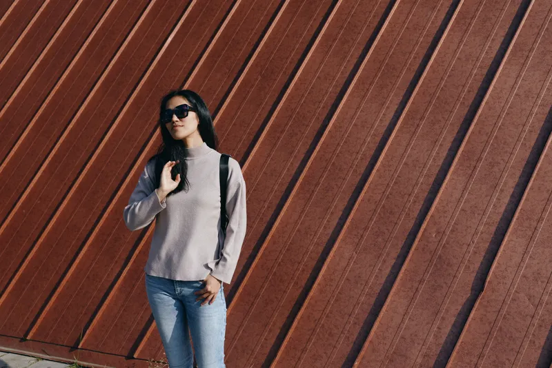 Fashion, street, model, sunglasses for urban style and casual outfit with backpack against a diagonal wooden wall. Woman in sweater and jeans poses confidently in daylight, showcasing contemporary