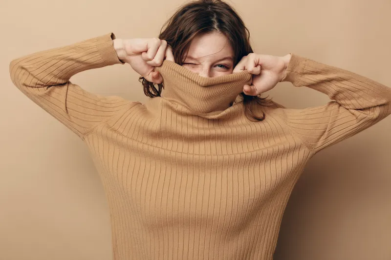 Young woman in beige knitted sweater pulls collar over her face, smiling with playful eyes on beige background. Concept of cozy fashion, autumn style, and casual comfort.