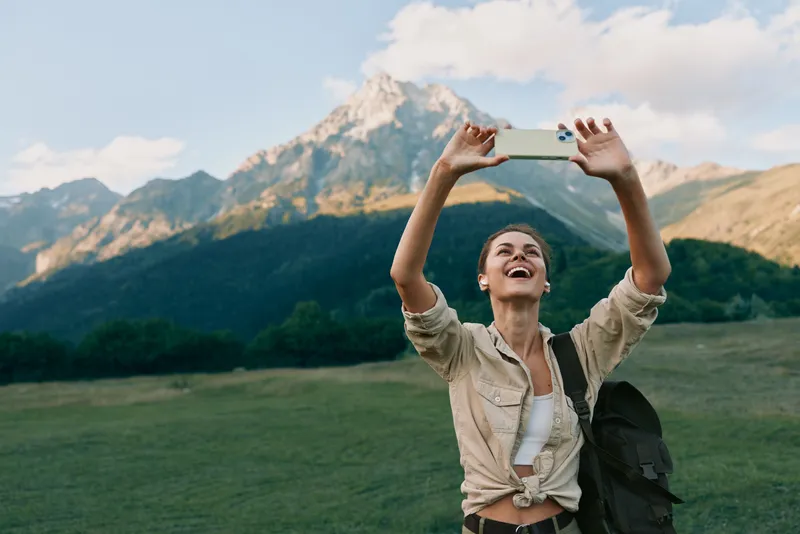 Young woman with a backpack smiles broadly while taking a selfie in a lush mountain meadow, capturing a moment of outdoor exploration and carefree fun.