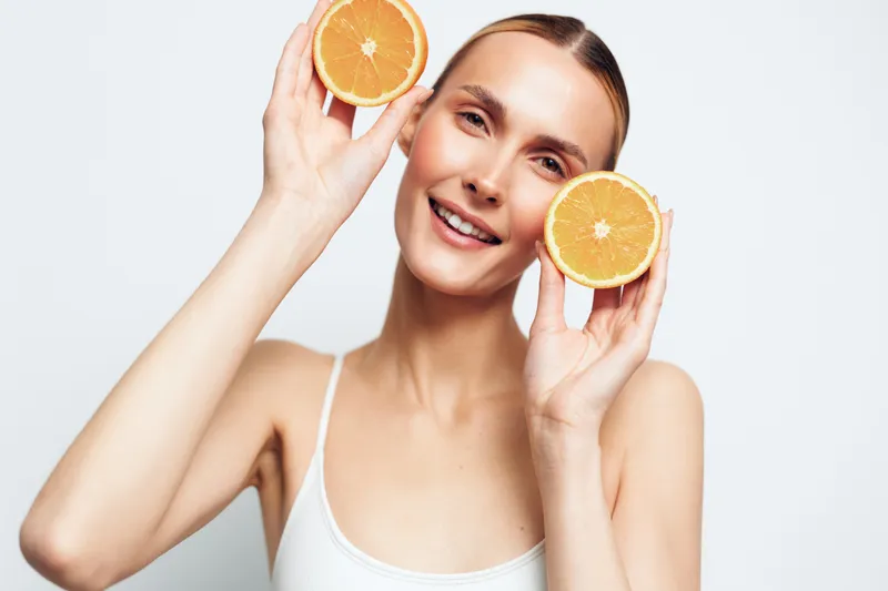 Smiling woman holding orange slices near her face on white background, fresh citrus beauty concept, natural glow skincare style, bright light, clean minimal mood, summer wellness care