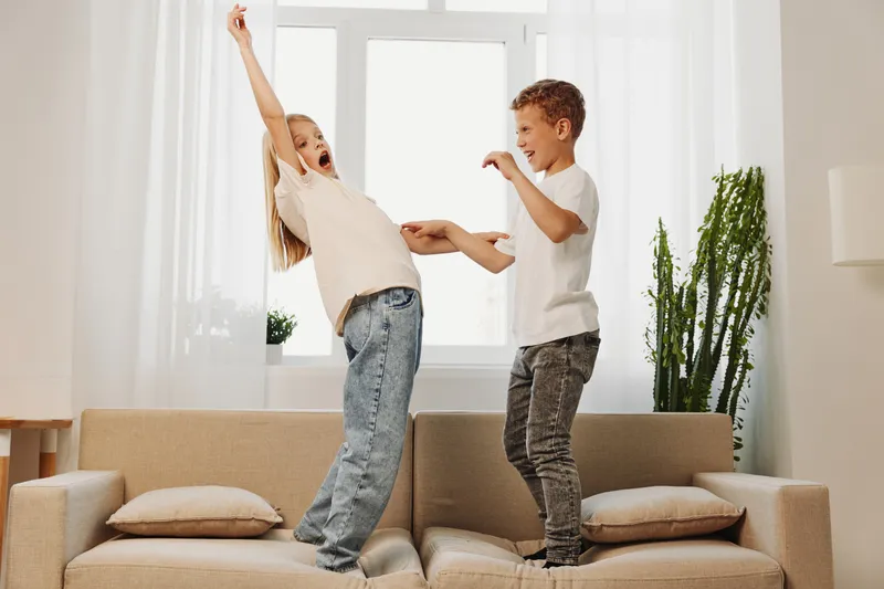 Two happy children standing on a beige sofa in bright living room playing and laughing. Casual clothes, natural light, and home environment create playful family fun concept.