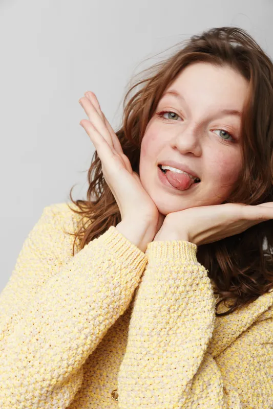 Young woman with brown hair wearing yellow sweater makes playful face with tongue out and hands near face. Studio portrait on light background. Concept of fun, youth, and casual mood.