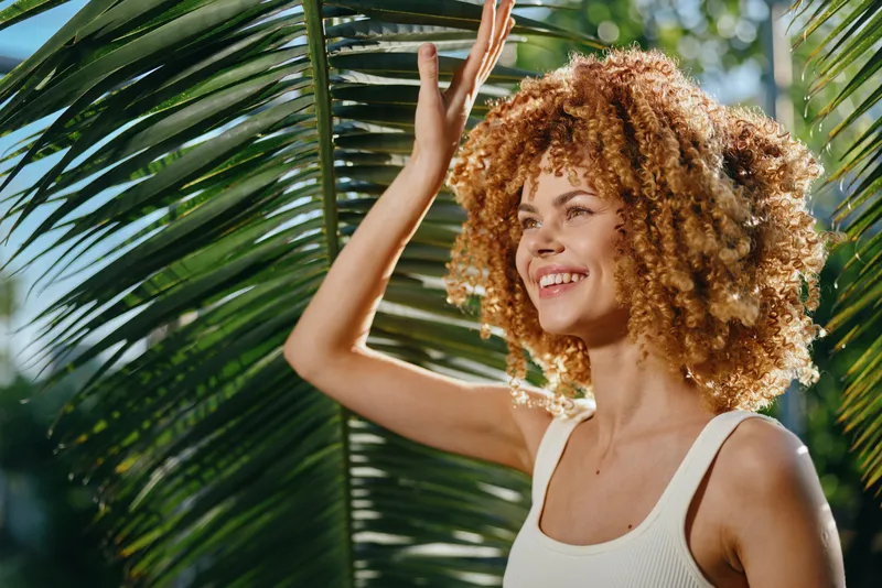 smiling woman with curly hair enjoying tropical sunshine outdoors near palm leaves, wearing white sleeveless top, natural light, vibrant green background