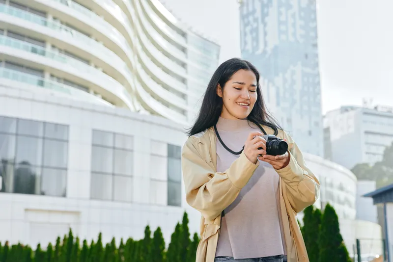 Woman with camera in urban city setting, smiling while photographing modern buildings and greenery, capturing outdoor travel and daily life through street level photography