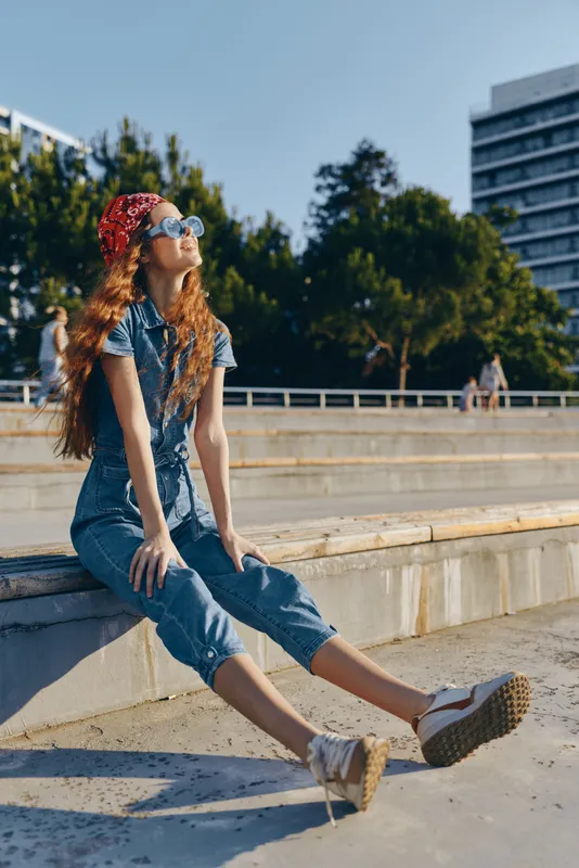 Lifestyle woman in modern boho-western denim street style with sunglasses and red bandana, enjoying sunny urban outdoor setting in film color aesthetic.