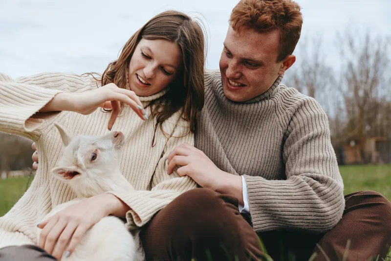 Happy couple outdoors with a playful goat kid, warm sweaters, gentle interaction, serene rural scene, lifestyle moment beside grass and clear sky, tender connection