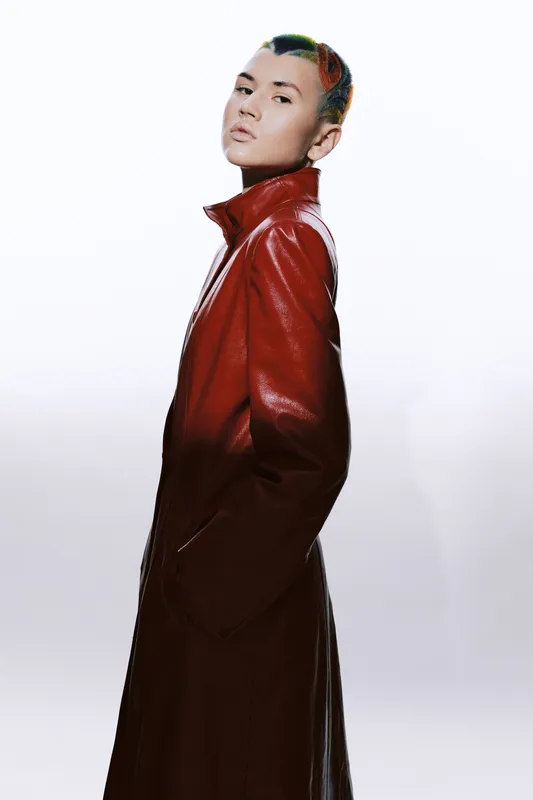 woman in long red leather coat posing confidently with hands in pockets, minimalistic white background, stylish fashion portrait with modern edgy look