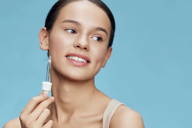 Young woman applying serum on her face with a dropper, showcasing a natural skincare routine against a light blue background, highlighting healthy skin and beauty
