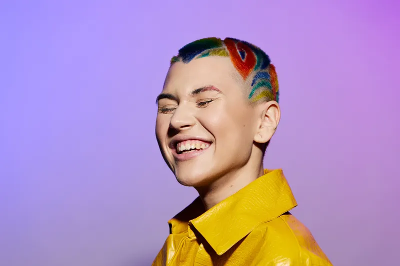 Smiling woman with colorful short hairstyle wearing a yellow jacket laughing joyfully against a purple background, expressing happiness and vibrant personality.