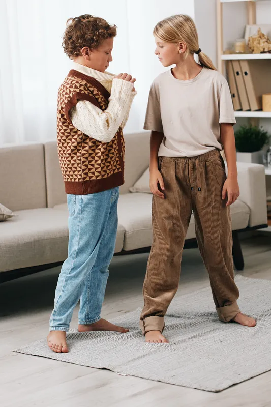 Two children stand on a soft rug indoors, facing each other with tense expressions, wearing casual cozy outfits, suggesting a quiet moment of conflict or conversation.