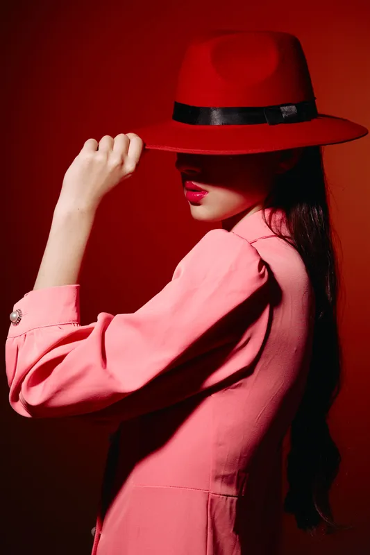 Fashionable woman wears red hat and pink jacket against red background. Her eyes are hidden under the hat while she lightly holds the brim. Concept for bold beauty and style.