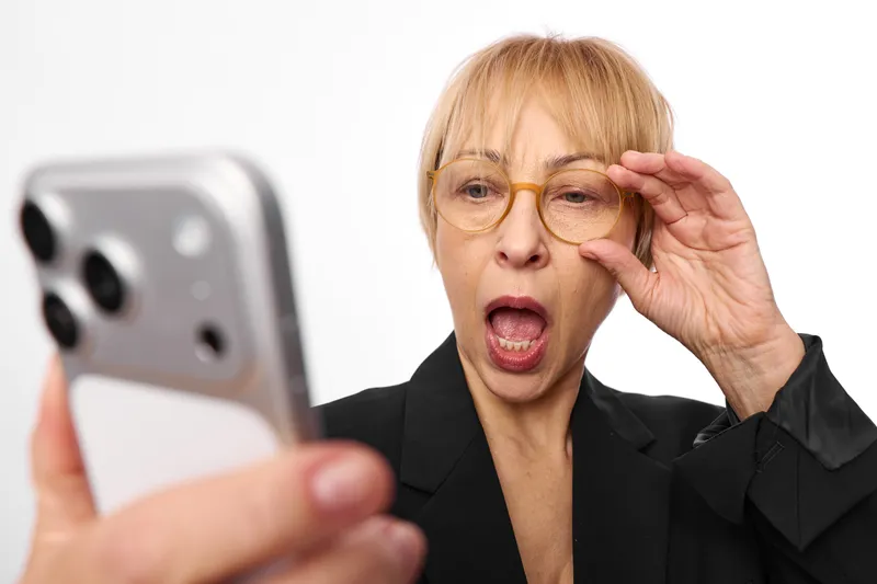 Elderly woman smartphone glasses business surprised concern hand indoors white portrait, executive holds mobile phone, adjusts eyewear, shocked expression, clean white background