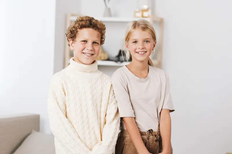 Two smiling children stand side by side indoors, wearing cozy sweaters in a bright, minimal living space. Soft natural light creates warm, inviting family vibes.