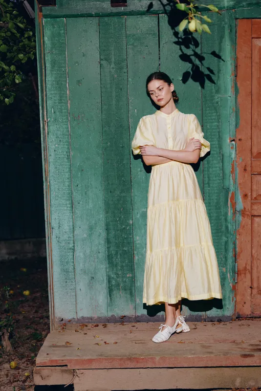 Woman in a pale yellow dress stands beside a green wooden doorway in an outdoor setting, a poised portrait with soft evening lighting, calm expression and flowing garment