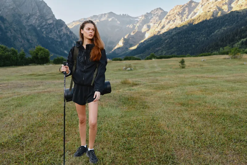 A cheerful explorer stands on an open meadow surrounded by rugged peaks. She holds a trekking pole, wears a dark jacket and shorts, and gazes ahead with a smile, radiating energy, confidence, and a