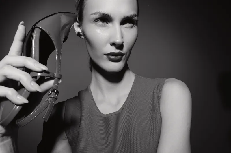 Portrait of a young white woman holding a high heel shoe near her face. Black and white studio photo with confident expression and stylish elegant look.